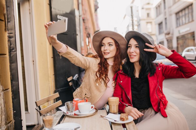 Fascinante mujer de jengibre haciendo selfie en café al aire libre con su amiga