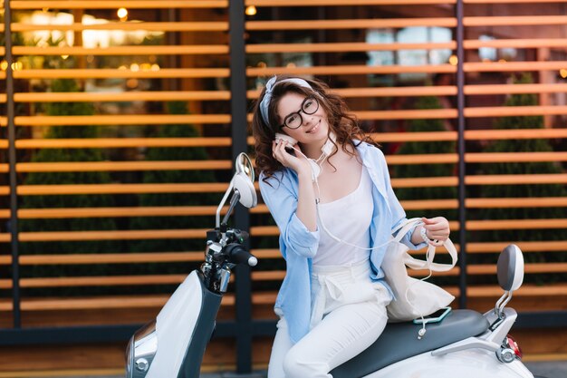 Fascinante señorita con elegantes gafas sosteniendo una bolsa ecológica blanca y escuchando música en auriculares, descansando después de comprar