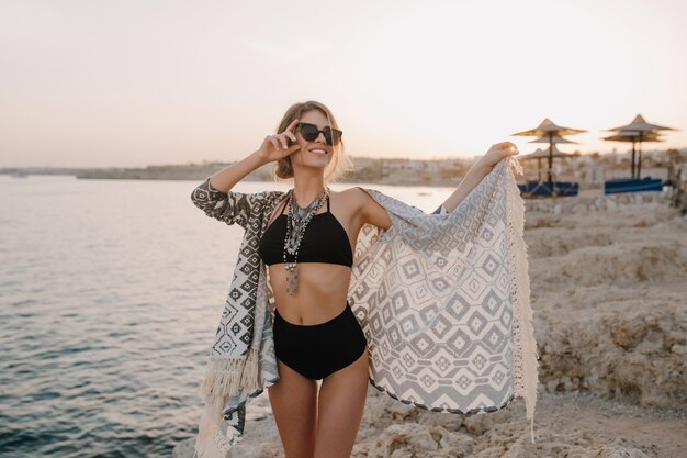 Fasionable hermosa chica posando en la playa, al atardecer. Mujer delgada sexy con bikini negro, traje de baño con cintura alta, chaqueta de punto, capa con adornos, hermoso mar, roca.