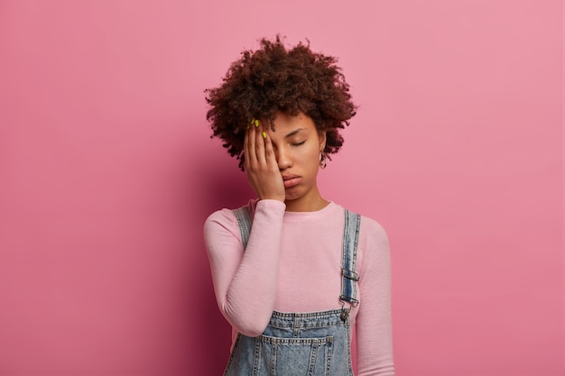 Fatiga La mujer de cabello rizado se siente aburrida y angustiada, quiere dormir, se cubre la mitad de la cara con la palma, mantiene los ojos cerrados, usa ropa de moda, posa contra la pared rosa. Concepto de cansancio