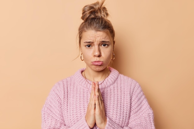 Por favor, ayúdame. Mujer joven molesta con monederos de pelo peinado lis mira tristemente a la cámara mantiene las palmas juntas te ruega que pidas perdón usa un suéter de punto casual aislado en un fondo beige