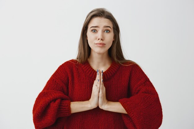 Por favor cariño, quiero anillo. Atractiva mujer caucásica con suéter rojo suelto, tomados de las manos en oración y levantando las cejas, pidiendo disculpas o pidiendo ayuda con la expresión de ángel y los ojos de cachorro