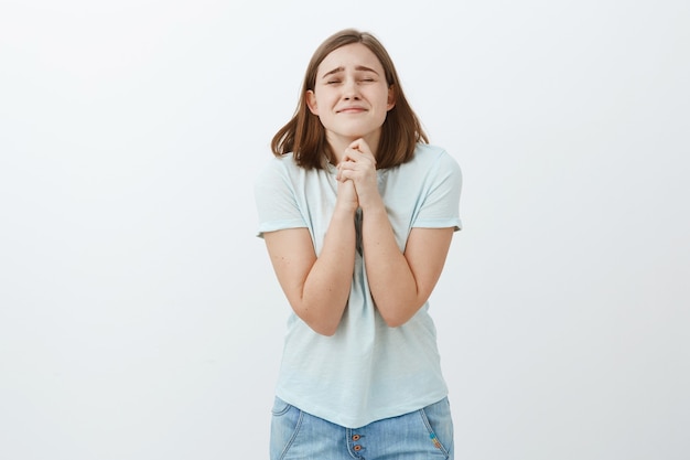 Por favor, hágalo realidad. Esperanza optimista y fiel estudiante linda cerrando los ojos y frunciendo los labios apretando las manos juntas cerca del cuerpo mientras pide un deseo o reza sobre una pared blanca