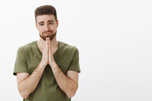 Por favor, hazme un favor, linda. Encantador, gentil y guapo carismático joven con barba y ojos azules haciendo una cara encantadora como suplicando pidiendo disculpas con las manos en oración sobre el pecho, sonriendo