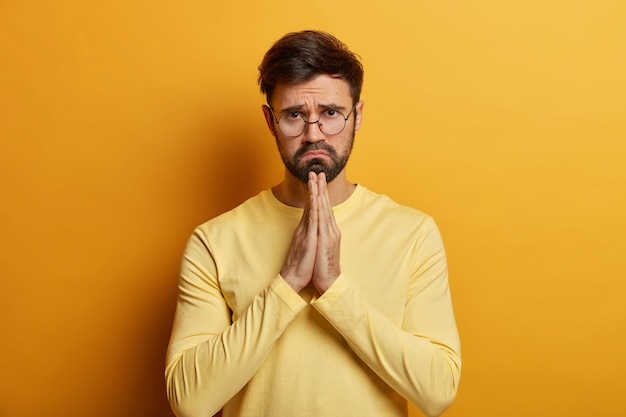 Por favor perdoname. Hombre deprimido sin afeitar aprieta las palmas de las manos y hace disculparse, tiene mirada inocente, pide ayuda, usa anteojos, suéter amarillo, mira con cara de lástima. La oración masculina pide hacer un favor