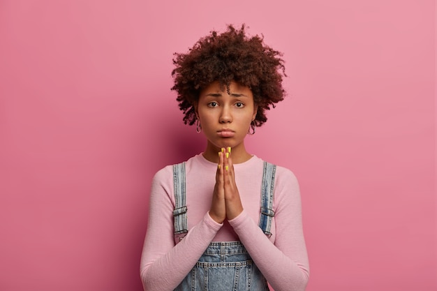 Por favor, te lo ruego. Triste mujer étnica de piel oscura toma las manos en oración, pide disculpas o ayuda, se frunce el labio inferior, tiene un peinado rizado, usa un cuello alto informal, le ruega a un amigo que la perdone