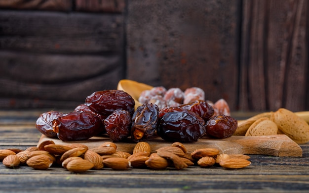 Fechas en una tabla de cortar con almendras peladas y sin pelar, nueces en vista lateral de cuchara de madera sobre fondo de azulejos de madera y piedra