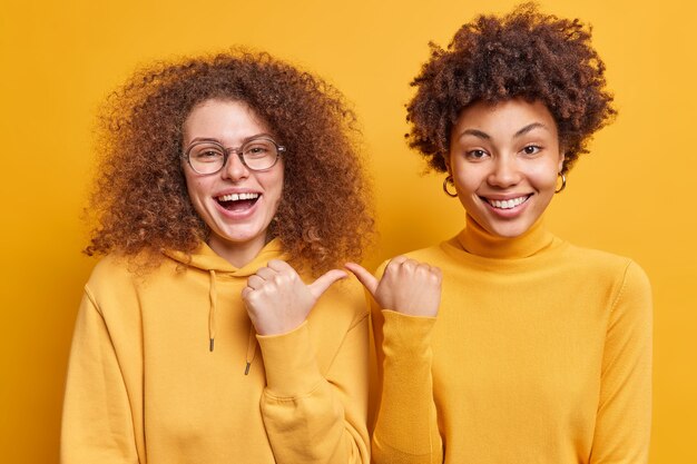 Felices y alegres mujeres de pelo rizado de raza mixta que se señalan entre sí con una expresión alegre y dicen que están de pie muy cerca una de la otra vestida informalmente aislada sobre una pared amarilla. Te escojo a ti