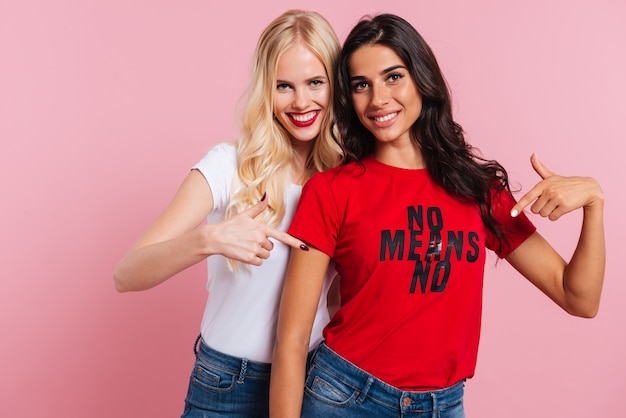 Felices amigas apuntando a la camisa con frase y sonriendo aislado