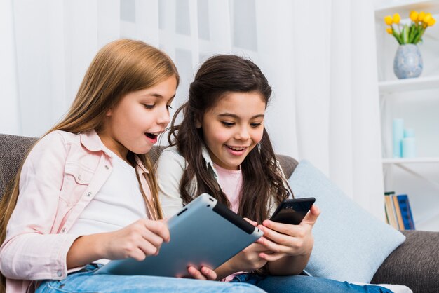 Felices amigas sentadas en el sofá con tableta digital y teléfono móvil