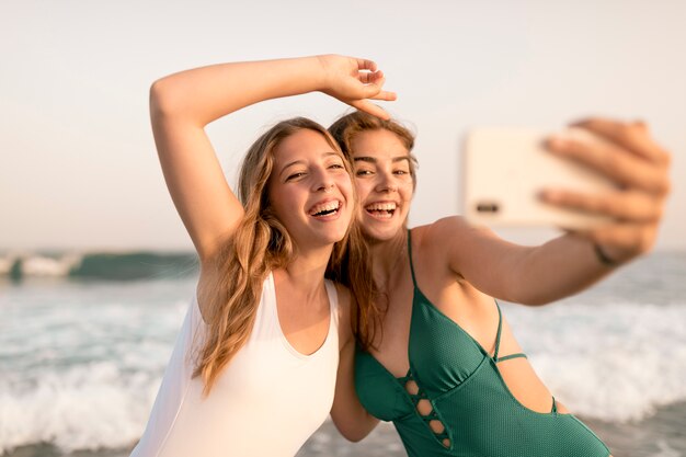 Felices amigas tomando selfie de teléfono inteligente a orilla del mar