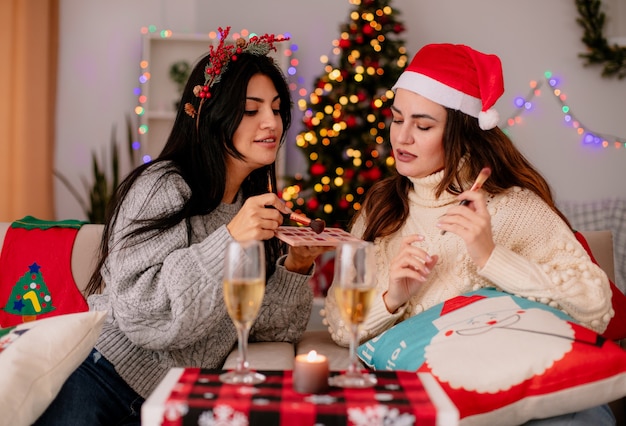 Felices chicas guapas sostienen pinceles en polvo haciendo maquillaje sentado en sillones y disfrutando de la Navidad en casa