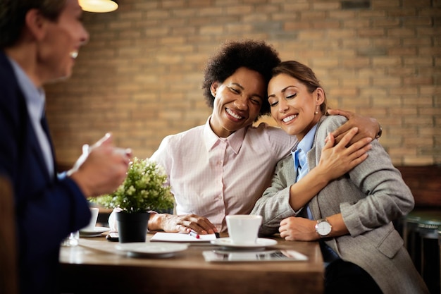 Felices colegas femeninas abrazándose con los ojos cerrados mientras toman un descanso para tomar café en un café