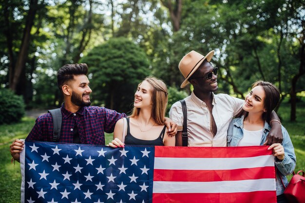 Felices cuatro estudiantes relajándose en la naturaleza con la bandera americana, celebrando el 4 de julio