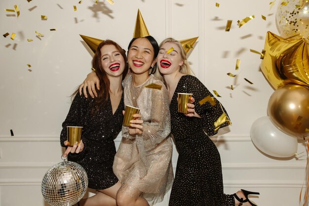Felices damas interraciales jóvenes en vestidos están caminando en la fiesta con globos de alcohol y confeti sobre fondo blanco Concepto de feliz fin de semana