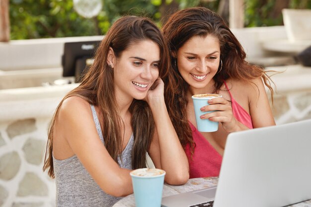 Felices dos hermanas hacen compras en línea, eligen un nuevo atuendo en Internet, miran alegremente la computadora portátil, beben café con leche o café