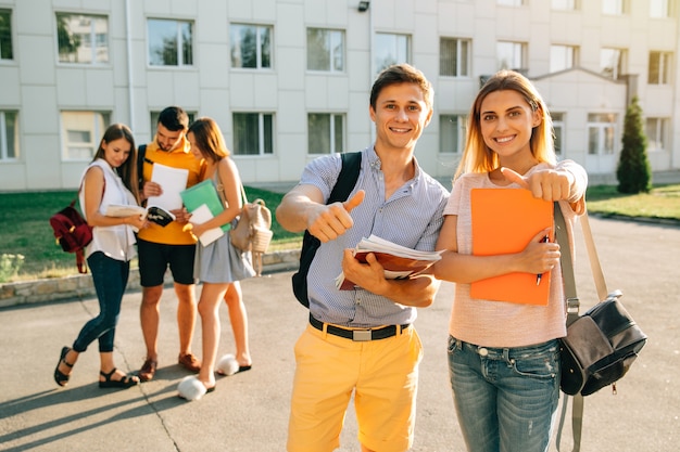 Felices dos jóvenes estudiantes con cuadernos y mochilas sonriendo y mostrando el pulgar hacia arriba