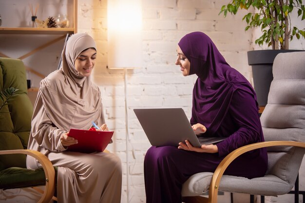 Felices dos mujeres musulmanas en casa durante la lección, estudiando cerca de la computadora, educación en línea