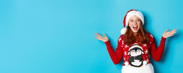 Felices fiestas concepto alegre pelirroja chica en suéter de navidad y gorro de santa levantando las manos y deseando