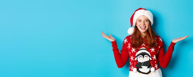 Foto gratuita felices fiestas y concepto de navidad alegre chica pelirroja con gorro de santa y suéter de navidad levantando han