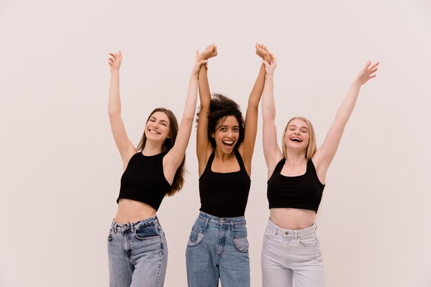 Felices jóvenes caucásicas y africanas con ropa informal divirtiéndose tomándose de la mano sobre sus cabezas con fondo blanco