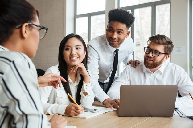 Felices jóvenes colegas de negocios usando la computadora portátil hablando entre sí.