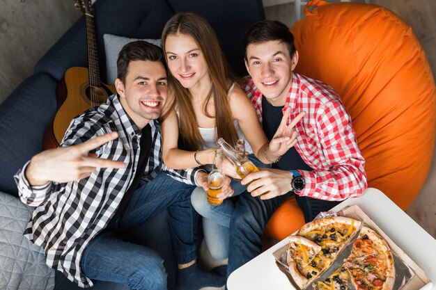 Felices los jóvenes comiendo pizza, bebiendo cerveza, brindando, divirtiéndose, fiesta de amigos en casa, compañía hipster juntos, dos hombres una mujer, sonriendo, positivo, posando para la foto