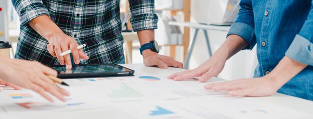 Felices jóvenes empresarios de Asia y mujeres de negocios que se reúnen para intercambiar ideas sobre nuevos colegas del proyecto de papeleo que trabajan juntos en la planificación de la estrategia de éxito disfrutan del trabajo en equipo en la pequeña oficina moderna.