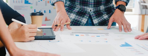 Felices jóvenes empresarios de Asia y mujeres de negocios que se reúnen para intercambiar ideas sobre nuevos colegas del proyecto de papeleo que trabajan juntos en la planificación de la estrategia de éxito disfrutan del trabajo en equipo en la pequeña oficina moderna.