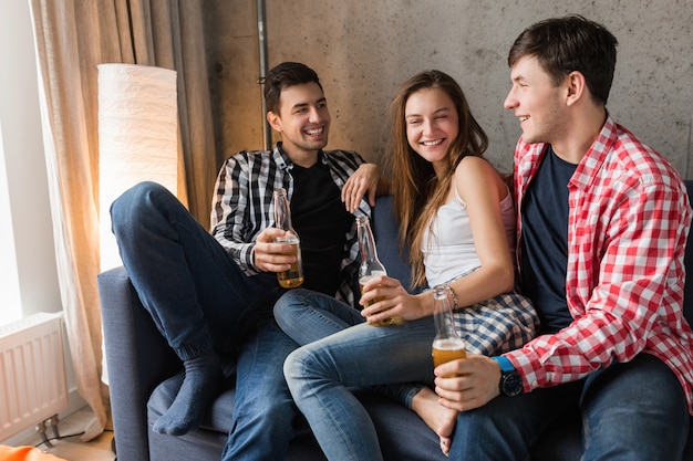 Foto gratuita felices los jóvenes sentados en el sofá, bebiendo cerveza, cerca de las manos brindando, divirtiéndose, fiesta en casa de amigos, compañía hipster juntos, dos hombres una mujer, sonriendo, positivo, relajado, pasar el rato, riendo