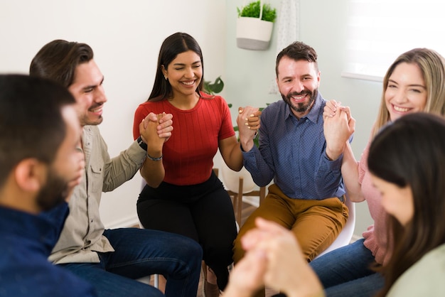 Felices miembros de un grupo de rehabilitación riéndose mientras se toman de la mano. Hombres y mujeres jóvenes alegres y emocionados en un grupo de apoyo