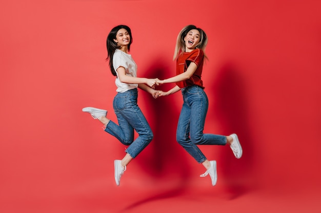 Felices mujeres asiáticas en elegantes camisetas y pantalones vaqueros saltando en la pared aislada