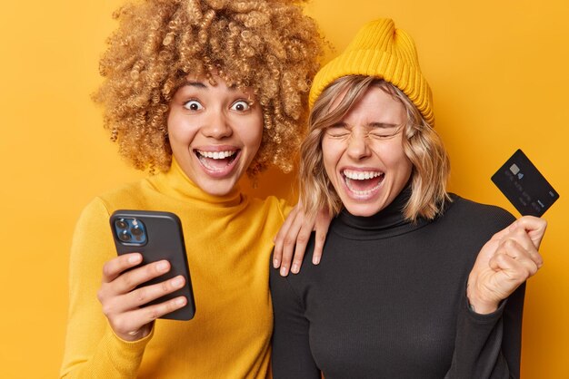 Felices mujeres jóvenes y amigables se ríen alegremente usan teléfonos móviles modernos y tarjetas de crédito para pagar en línea contentas de gastar dinero en compras paradas una al lado de la otra aisladas sobre fondo amarillo