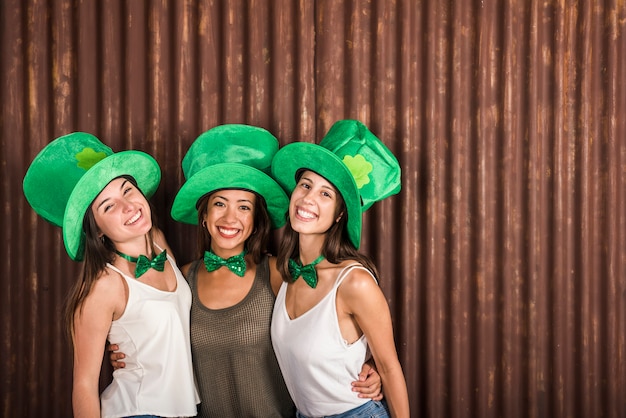 Felices mujeres jóvenes con sombreros de San Patricio abrazando cerca de la pared