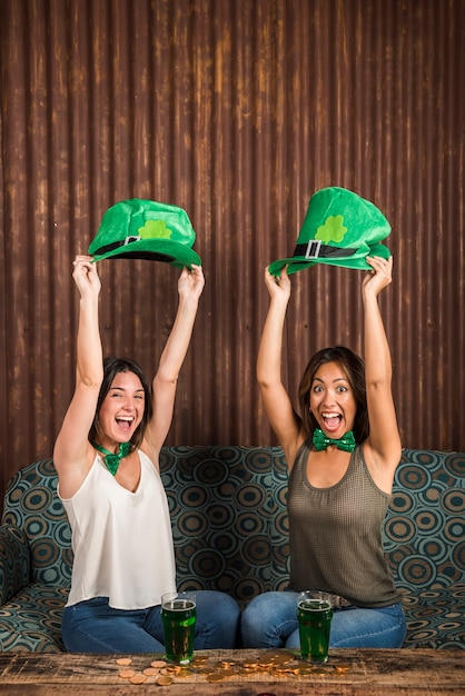 Foto gratuita felices mujeres jóvenes con sombreros de san patricio cerca de la mesa con vasos de bebida