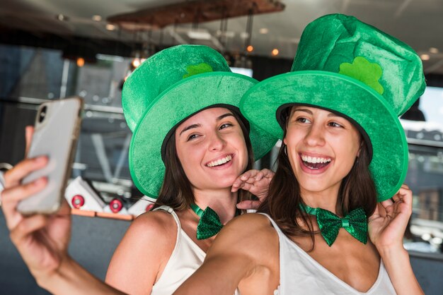 Felices mujeres jóvenes en sombreros de San Patricio que toman selfie en teléfono inteligente