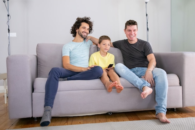 Felices padres homosexuales e hijo sentados juntos en el sofá en casa y viendo comedia en la televisión, mirando a otro lado, sonriendo y riendo. Concepto de familia y paternidad