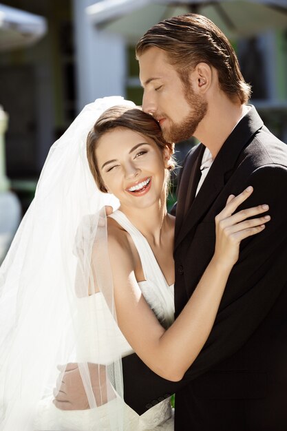 Felices recién casados hermosos sonriendo, abrazándose, besándose afuera.