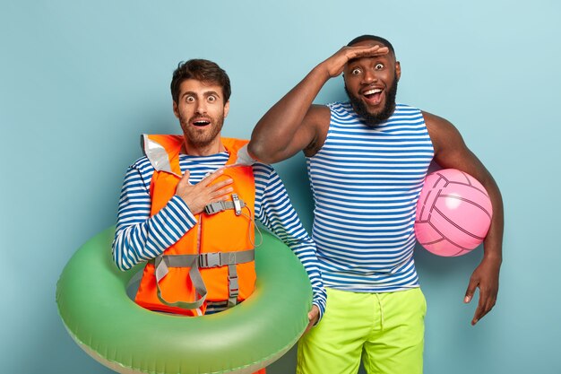 Felices sorprendidos hombres jóvenes diversos vestidos con pantalones cortos y camiseta de marinero, sostienen la pelota de playa