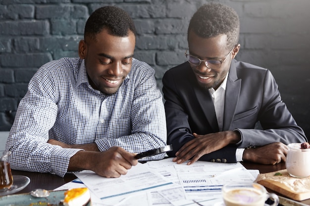 Felices trabajadores de oficina afroamericanos vestidos con ropa formal con una apariencia alegre, estudiando y asombrando documentos legales en la mesa con lupa mientras preparan papeles para la reunión