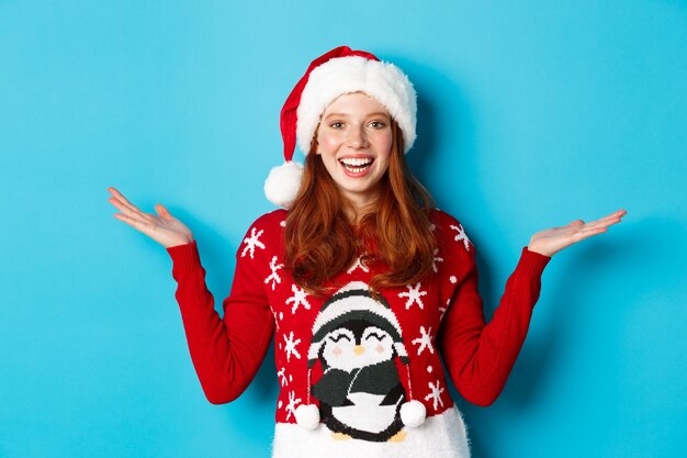 Felices vacaciones y concepto de Navidad. Chica pelirroja alegre con gorro de Papá Noel y suéter de Navidad, levantando las manos en los espacios de copia, sosteniendo algo sobre fondo azul.