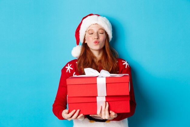 Felices vacaciones y concepto de Navidad. Linda chica pelirroja con regalos y labios fruncidos para besar, vistiendo gorro de Papá Noel y suéter divertido, fondo azul.