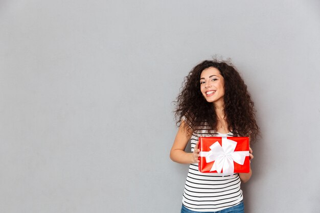 Felicidad y alegría expresando joven sosteniendo caja envuelta para regalo con lazo blanco mientras está de pie sobre la pared gris