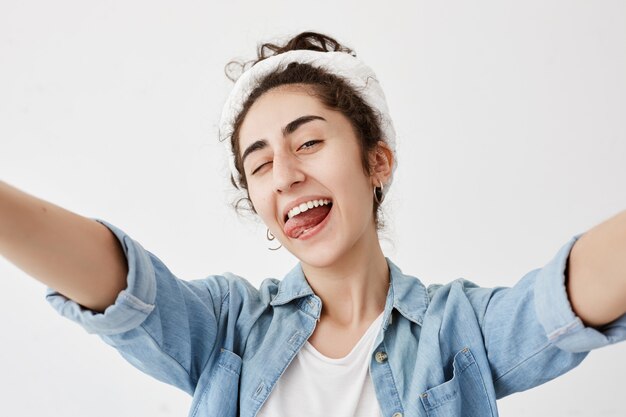 Felicidad, belleza, alegría y juventud. Joven positiva vestida con camisa vaquera sobre camiseta blanca estirando los brazos, sonriendo ampliamente, parpadeando, sacando la lengua, de buen humor.