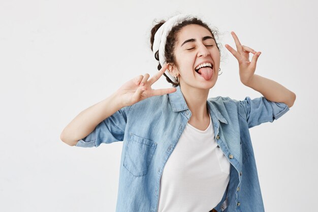 Felicidad, belleza, alegría y juventud. Joven positiva vestida con camisa vaquera sobre camiseta blanca que muestra el signo v, sonriendo ampliamente, con los ojos cerrados, sacando la lengua, de buen humor.