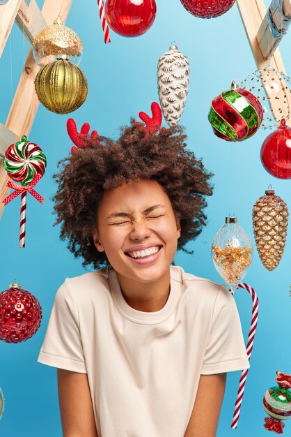 Felicidad y concepto de evento festivo. Alegre alegre mujer de piel oscura se ríe, cierra los ojos y sonríe ampliamente, va a decorar el árbol de Navidad, usa una camiseta blanca informal, disfruta de las vacaciones de invierno
