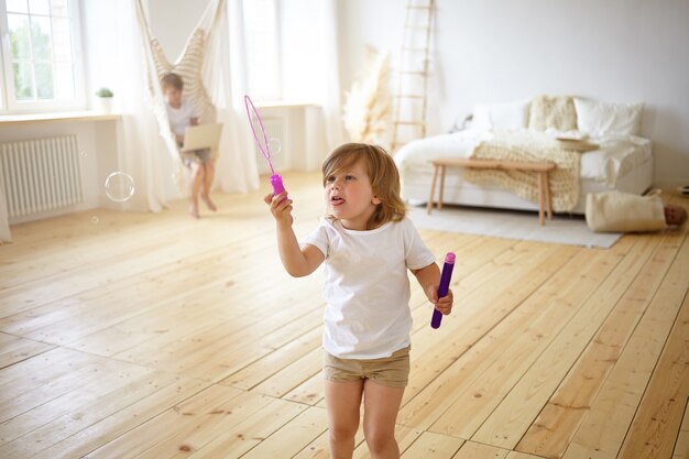 Felicidad y concepto de infancia sin preocupaciones. Retrato interior de adorable niña feliz en camiseta y pantalones cortos de pie en medio de la moderna sala de estar, divirtiéndose, soplando pompas de jabón,