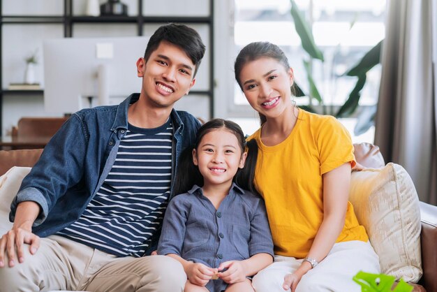 Felicidad, familia asiática joven, papá, mamá e hija, informales, relajados, sentados, sonriendo, pasando un momento de paz juntos en el sofá de la sala de estar en casa.