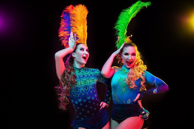 Foto gratuita felicidad. hermosas mujeres jóvenes en carnaval, elegante disfraz de mascarada con plumas sobre fondo negro en luz de neón. copyspace para anuncio. celebración de fiestas, baile, moda. tiempo festivo, fiesta.