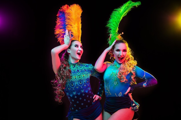 Felicidad. Hermosas mujeres jóvenes en carnaval, elegante disfraz de mascarada con plumas sobre fondo negro en luz de neón. Copyspace para anuncio. Celebración de fiestas, baile, moda. Tiempo festivo, fiesta.